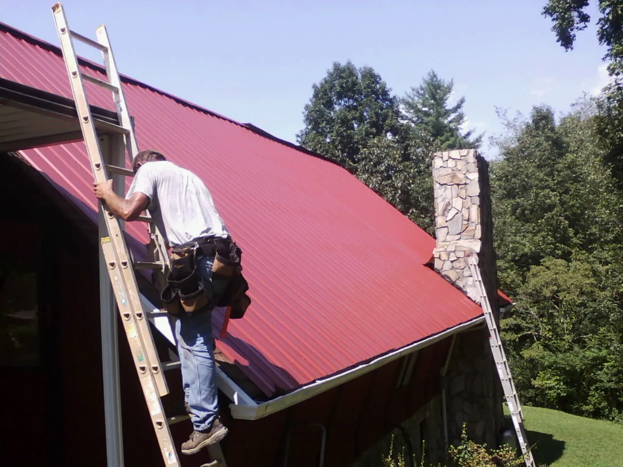 Roofer on a ladder beside a red metal roof and stone chimney — Soffit & Fascia Repair by Roofing Brothers in Dunmor